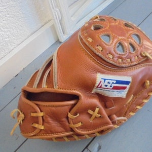 Puede incluir: Un guante de receptor de béisbol de cuero marrón con costuras blancas y un logotipo "NSG" blanco, rojo y azul.