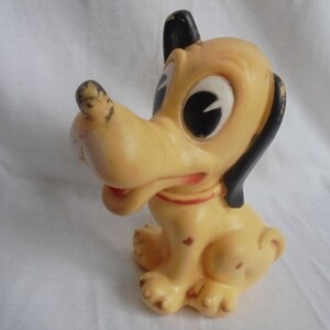 May include: A vintage yellow rubber toy dog with black ears and large black eyes. The dog is sitting with its tongue sticking out and has a red collar.