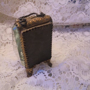 May include: A small, ornate, gold-toned picture frame with a beveled glass front and a dark brown backing. The frame has a decorative design and stands on four small feet.