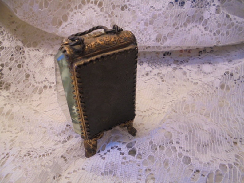 May include: A small, ornate, gold-toned picture frame with a beveled glass front and a dark brown backing. The frame has a decorative design and stands on four small feet.