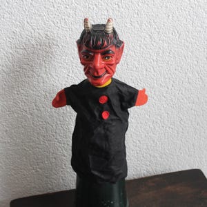 May include: A hand puppet featuring a molded red devil head with small white horns, pointed ears, and a black painted hairstyle. The puppet has a black fabric body with two red circular buttons on the front and red fabric hands extending from the sleeves.