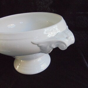 May include: White porcelain serving bowl with two ornate handles. The bowl has a wide, shallow base and a rounded, slightly flared rim.