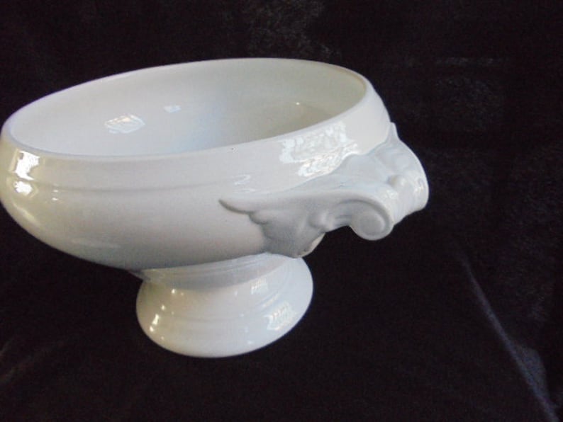 May include: White porcelain serving bowl with two ornate handles. The bowl has a wide, shallow base and a rounded, slightly flared rim.