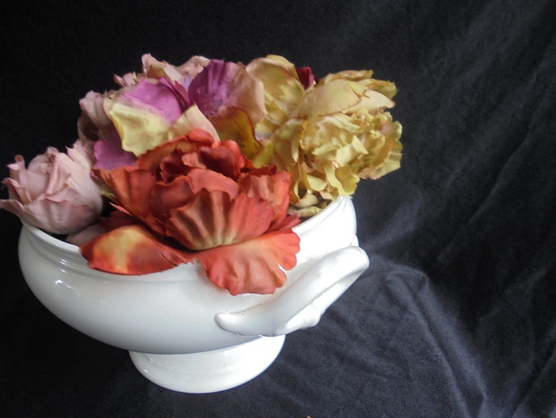 May include: A white ceramic bowl filled with a variety of artificial flowers, including pink, yellow, and orange roses.