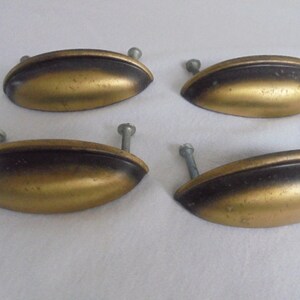 Op de afbeelding: Vier messingkleurige metalen kastgrepen met een zwarte afwerking. Elke greep heeft een afgeronde ovale vorm met een lichte bocht en twee zilveren schroeven.