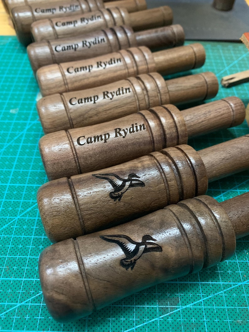 Op de afbeelding: Een verzameling houten eendenlokkers met de tekst "Camp Rydin" gegraveerd. Sommige lokkers hebben een gedetailleerde zwarte illustratie van een eend. De lokkers zijn gemaakt van donkerbruin hout.