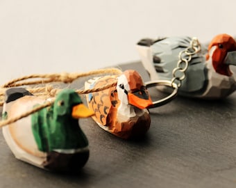 May include: Hand-carved wooden duck figurines in green, brown, orange, and gray, with orange beaks. One duck is attached to a keychain. The ducks are connected by a natural twine.