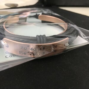 May include: A black leather bracelet with a rose gold band engraved with the coordinates "20&deg; 40' 49.2"N 156&deg; 26' 29.1"W".