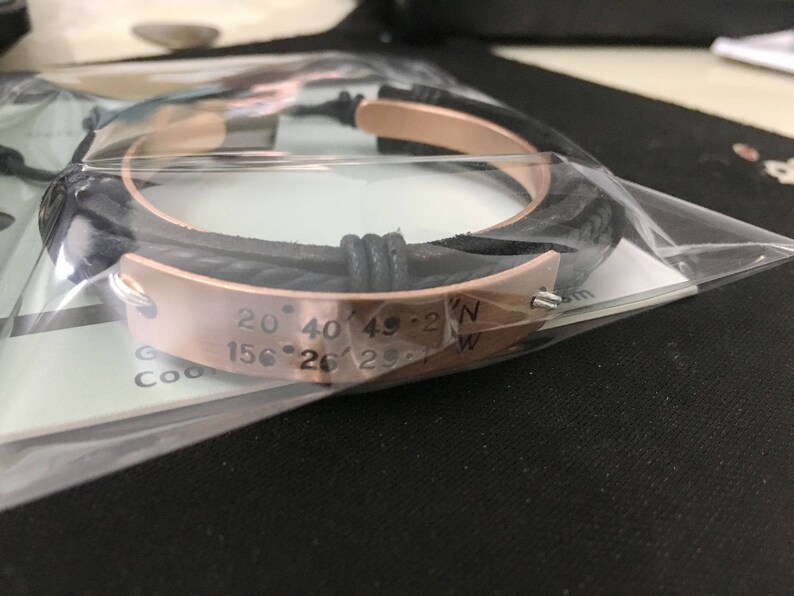 May include: A black leather bracelet with a rose gold band engraved with the coordinates "20&deg; 40' 49.2"N 156&deg; 26' 29.1"W".