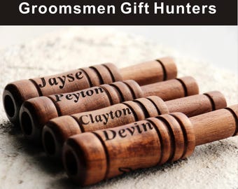 May include: Wooden duck calls with engraved names: Jayse, Peyton, Clayton, and Devin. The calls are brown with a dark finish and are arranged on a textured surface. The text "Groomsmen Gift Hunters" is at the top.