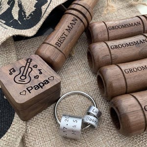 Op de afbeelding: Een verzameling gepersonaliseerde houten geschenken, waaronder een doos met een gitaar en het woord "Papa", en een sleutelhanger met gegraveerde ringen. Ook afgebeeld zijn houten fluitjes met de inscripties "Best Man" en "Groomsman".
