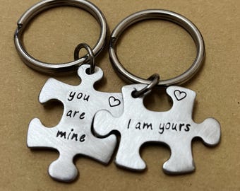 May include: Two silver puzzle piece keychains. One piece reads "you are mine" and the other reads "I am yours", each with a small heart. The keychains are attached to silver key rings.