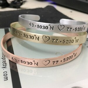 May include: Three metal bracelets with a heart and coordinates engraved on them. The bracelets are silver, gold, and copper.
