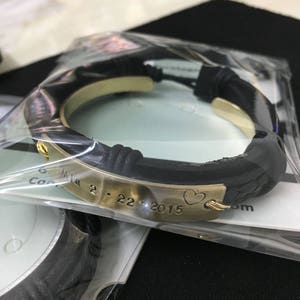 May include: A black leather bracelet with a gold metal band engraved with the date "2.22.2015" and a heart symbol.