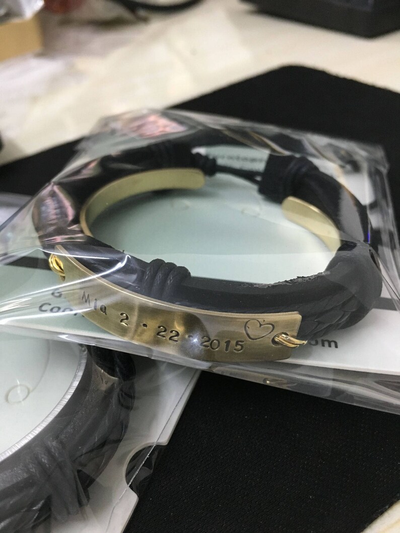 May include: A black leather bracelet with a gold metal band engraved with the date "2.22.2015" and a heart symbol.