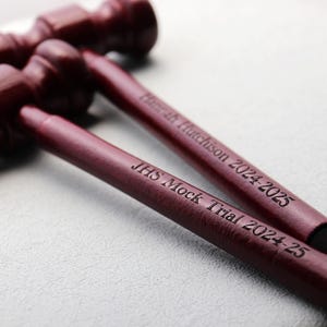 Pode incluir: Três martelos de madeira castanho-avermelhada dispostos diagonalmente. Os textos "JHS Mock Trial 2024-25" e "Hannah Hutchinson 2024-2025" estão gravados nos cabos. Os martelos estão sobre uma superfície cinza claro.