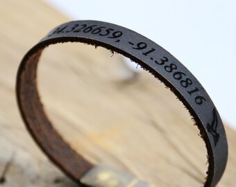 May include: A close-up of a leather bracelet with a dark gray band and a brown interior. The band is engraved with coordinates and a bird silhouette. The bracelet has a metallic clasp.