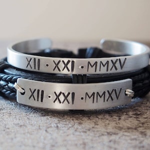 Può includere: Due braccialetti in pelle nera con bande in metallo argentato. Le bande sono incise con numeri romani e l'anno 2015. I braccialetti sono legati insieme da un cordoncino nero.