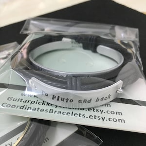 May include: A black leather bracelet with a silver metal band engraved with the text "To Pluto and back".
