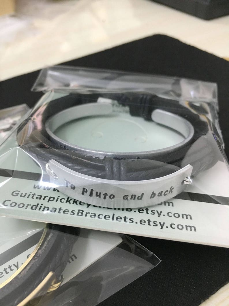 May include: A black leather bracelet with a silver metal band engraved with the text "To Pluto and back".