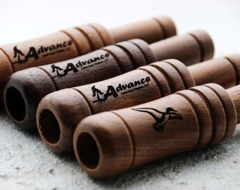 May include: A collection of wooden duck calls in varying shades of brown. Each call features the "Advance Land and Timber, LLC" logo, with some showcasing a tree graphic and others a flying duck silhouette. The calls are arranged on a textured surface.