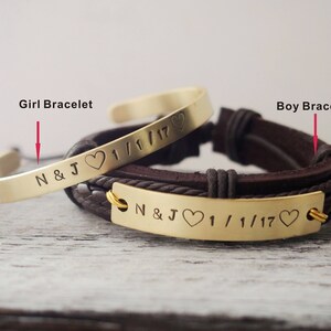 May include: Two gold bracelets with engraved text and hearts. The bracelet on the left is a simple bangle with the text "N&J 1/1/17". The bracelet on the right is a brown leather strap with a gold plate that reads "N&J 1/1/17".