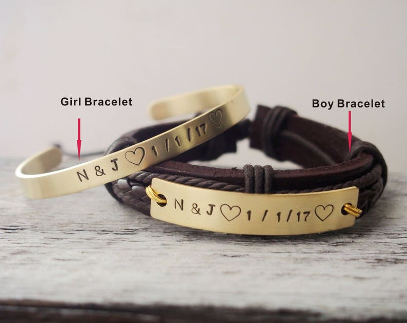 May include: Two gold bracelets with engraved text and hearts. The bracelet on the left is a simple bangle with the text "N&J 1/1/17". The bracelet on the right is a brown leather strap with a gold plate that reads "N&J 1/1/17".