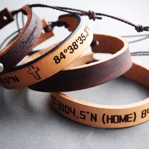 Può includere: Tre braccialetti in pelle con coordinate incise e una croce. I braccialetti sono marroni, beige e marrone chiaro. Il testo sui braccialetti recita "Deuteronomy", "84°38′35.3"", "4.5"N", "63104.5"N (HOME) 84°38′".