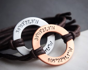 May include: Two personalized bracelets with circular metal pendants. One is silver, the other is copper-colored. Each pendant has engraved coordinates and is attached to a brown cord. The text on the silver bracelet reads "38°03'27.3"N". The copper bracelet reads "38°03'27.3"N" and "105°22'11.1"W".