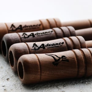 Op de afbeelding: Een verzameling houten eendenlokkers met een natuurlijke afwerking. Elke lokker heeft het "Advance Land And Timber, LLC" logo en een silhouet van een eend. De lokkers zijn diagonaal gerangschikt, wat hun cilindrische vorm en gegraveerde details benadrukt.