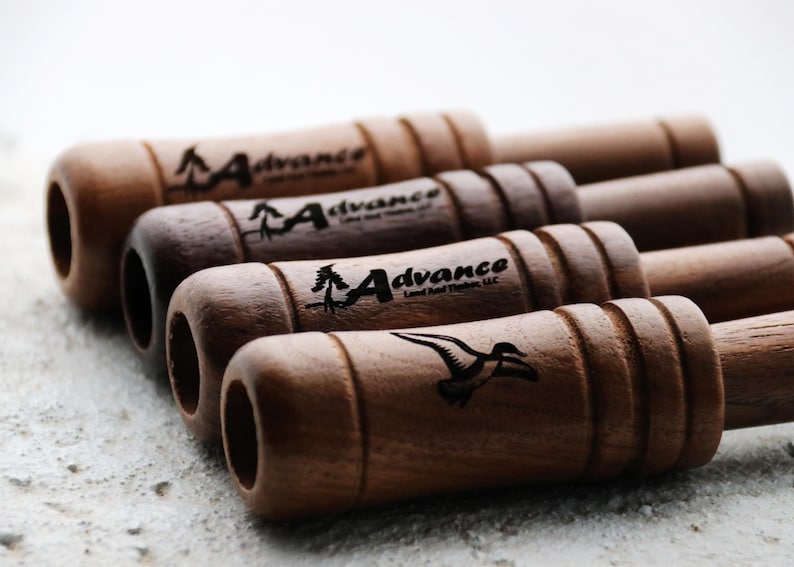 Op de afbeelding: Een verzameling houten eendenlokkers met een natuurlijke afwerking. Elke lokker heeft het "Advance Land And Timber, LLC" logo en een silhouet van een eend. De lokkers zijn diagonaal gerangschikt, wat hun cilindrische vorm en gegraveerde details benadrukt.