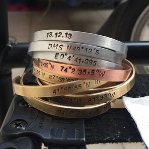 May include: A stack of six metal bracelets with engraved coordinates and dates. The bracelets are in different colors, including silver, copper, and brass.