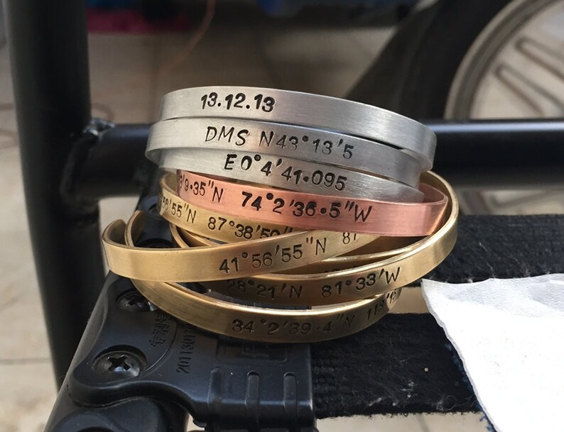 May include: A stack of six metal bracelets with engraved coordinates and dates. The bracelets are in different colors, including silver, copper, and brass.