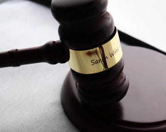 May include: A dark brown wooden gavel with a matching base. A gold band encircles the gavel head, with the inscription "Sarah Wins". The gavel is set against a light-colored background, suggesting a formal setting.