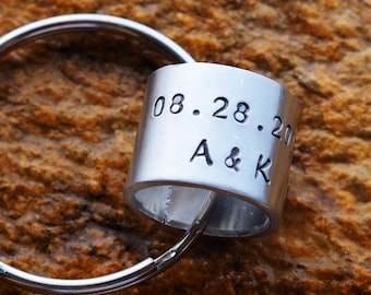 May include: A silver-colored keychain with a cylindrical tag. The tag is engraved with the date "08.28.20" and the initials "A & K". The keychain is set against a textured, brown background.