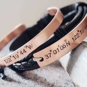 May include: Two copper bracelets with black leather straps. One bracelet has the coordinates "37&deg; 01' 05"N 122&deg; 19' 44"W" engraved on it. The other bracelet has the coordinates "122&deg; 19' 44"W" engraved on it.