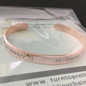May include: A rose gold bangle bracelet with the coordinates "73.989459&deg; W 40.729527&deg; N" engraved on the outside.