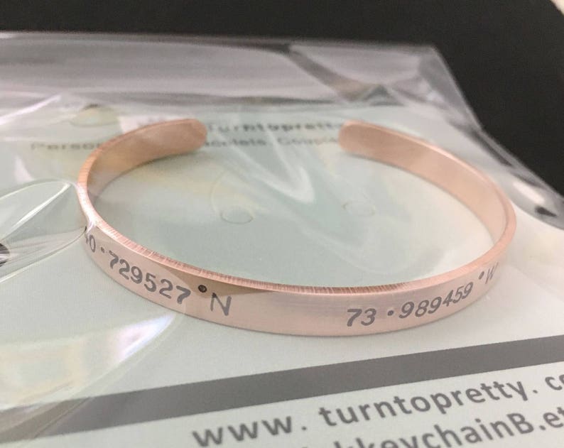 May include: A rose gold bangle bracelet with the coordinates "73.989459&deg; W 40.729527&deg; N" engraved on the outside.