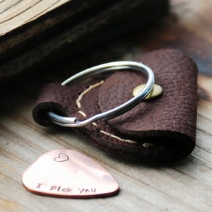 May include: A brown leather guitar pick holder with a silver key ring and a copper guitar pick that says "I Pick You" with a small heart.