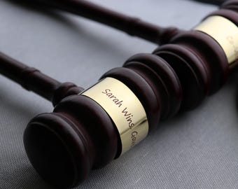 May include: Close-up of two wooden gavels with gold bands. The bands are engraved with the text "Sarah Wins, Gave". The gavels are dark brown and sit on a gray surface.