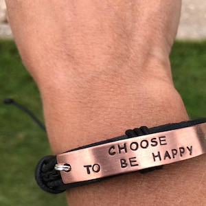 May include: A black leather bracelet with a copper plate that reads "Choose to be happy".