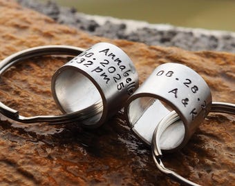 May include: Two silver keychains with engraved cylindrical charms. One charm reads "Annabella 08.2025 4:32 pm". The other charm reads "08.28 A & K". The keychains are on a textured brown surface.