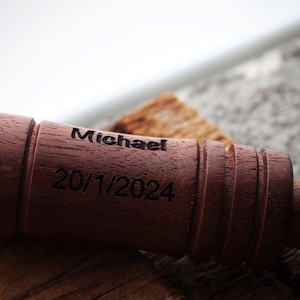 Op de afbeelding: Een close-up van een donkerbruin houten object met de naam "Michael" en de datum "20/1/2024" gegraveerd. Het object heeft een cilindrische vorm met een geribbeld ontwerp aan &eacute;&eacute;n uiteinde.