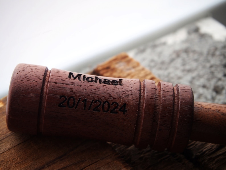 Op de afbeelding: Een close-up van een donkerbruin houten object met de naam "Michael" en de datum "20/1/2024" gegraveerd. Het object heeft een cilindrische vorm met een geribbeld ontwerp aan &eacute;&eacute;n uiteinde.