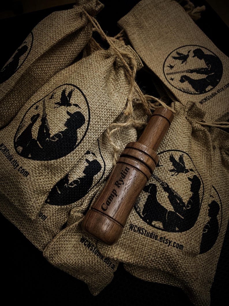 Op de afbeelding: Een houten eendenlokker met de tekst "Camp Rydin" ligt op een jute zak. De zak heeft een zwarte afbeelding van een jager en hond met vliegende eenden. Verschillende soortgelijke zakken zijn op de achtergrond.