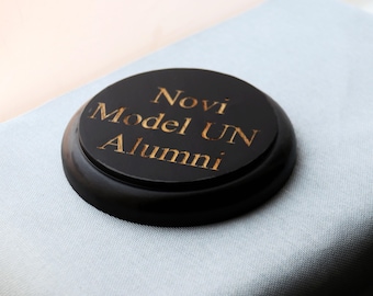 May include: A circular black wooden plaque with a beveled edge. The center features gold-colored engraved text that reads Novi Model UN Alumni. The plaque has a smooth, polished finish and is presented as a commemorative or award-style item.