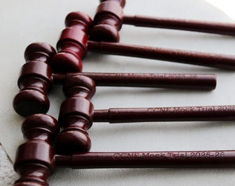 May include: Several dark red wooden gavels are arranged diagonally. The handles of the gavels have the text "CCNE Mock Trial 2025-26" engraved on them. The gavels are likely used in a courtroom or mock trial setting.