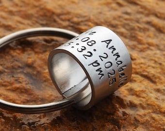 May include: A silver-colored ring with engraved text, attached to a silver key ring. The ring is inscribed with the name "Annabelle", the date "08.2025", and the time "5:32 pm". The ring is displayed against a textured, brown surface.