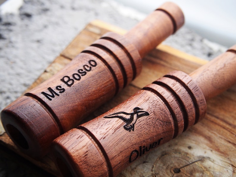 Op de afbeelding: Twee houten eendenlokfluiten met gegraveerde details. Op &eacute;&eacute;n staat "Ms Bosco", op de andere "Oliver" en een eendenafbeelding. De lokfluiten zijn bruin van kleur en hebben een geribbeld ontwerp.