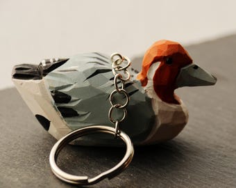 May include: A hand-carved wooden duck keychain. The duck has a red head, gray body, and white accents. A silver key ring and chain are attached to the duck's back. The keychain is designed to be a decorative accessory.